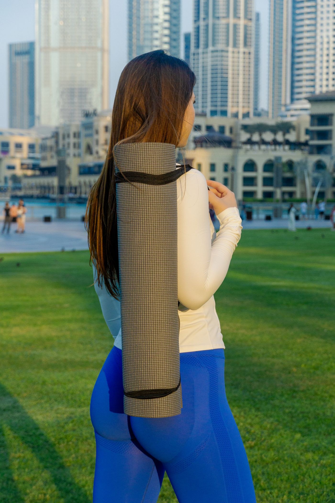 A woman carrying a rolled exercise mat over her shoulder in a park.