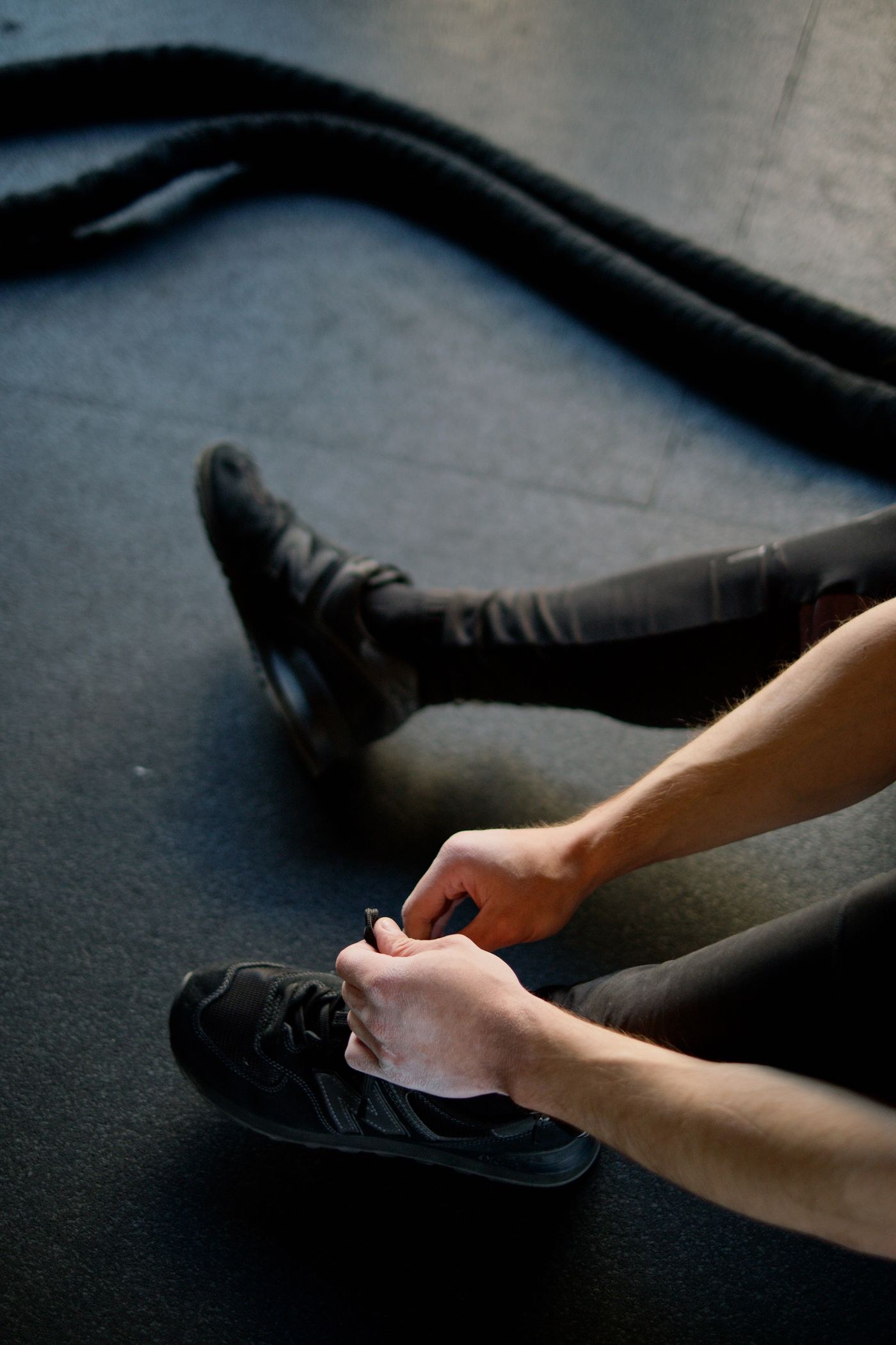 A person fastening black training shoes on a gym floor beside a battle rope.
